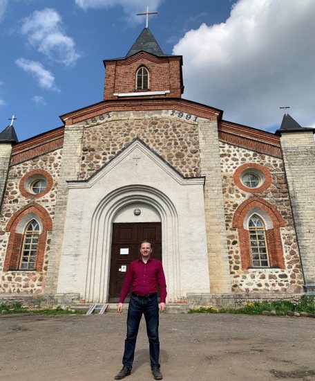 Исследователь Сергей Тамби перед евангелическо-лютеранской церковью Святого Иоанна Крестителя. В начале XX века службы в приходе Купанитса (фин. Kupanitsa) проводились на эстонском и финском языках. Деревня Губаницы Волосовского района Ленинградской области, 14 мая 2023 года. Источник: фотография из личного архива С. А. Тамби